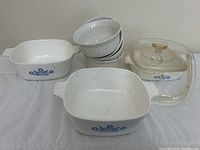 Set of white Corningware dishes showing square and oval casseroles with blue floral pattern, stacked white bowls, and glass lid