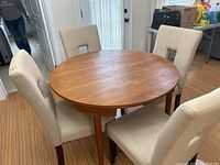 Full view of round wooden dining table with four beige upholstered chairs around it on wood flooring.