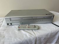 Philco 4-head DVD and VHS combo player with front panel and power cable with remote control in front.