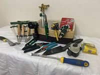 Wide view of assorted gardening tools arranged on white surface including wooden handled trowels, pruning shears, watering nozzle, sprinkler head, measuring tape, and yellow box