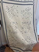 Full view of the single bed quilt hung vertically showing embroidered gray leaf designs and gray and ivory border stripes.