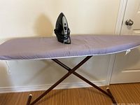 Wooden ironing board with light purple cover and folded crossed legs, photo taken to show the full ironing board.