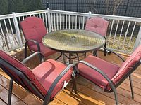 Outdoor round table with textured glass top and umbrella hole, surrounded by four cushioned red chairs on wood decking.