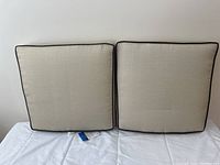 Two beige square porch cushions with dark brown piping laid flat against a white background