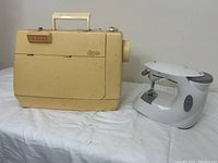 Side view of the vintage Singer Genie sewing machine in cream with handle on top and small handheld sewing machine next to it on white cloth background.