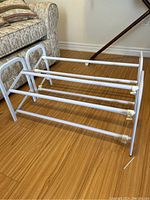 Two matching adjustable metal shoe racks on wood floor, showing tubular metal construction and curved end handles. Shoe racks are extended to current length.