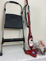 Photo showing electronic red mop with power cord, mop head, and attached folded step stool alongside it.