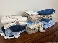 Stack of folded bedding showing multiple pillowcases, pillow shams, and sheets in various colors and patterns on a wooden surface
