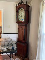 Full view of wooden longcase clock standing next to table with glassware