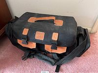 Closed black canvas camera bag with tan leather accents resting on carpet
