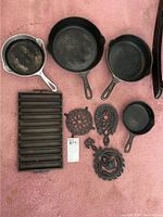 Overview of entire set including four frying pans, cornbread pan, and three decorative trivets on pink carpet showing condition with rust spots.