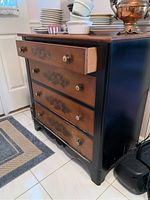 Side angle showing four drawer chest with black frame and wood tone drawer fronts having fruit transfer art and decorative knobs. Shows some wear and damage at bottom.