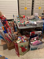 Wide view of lot on table and floor showing wrapping paper rolls in box, bins of ribbons and gift bags.
