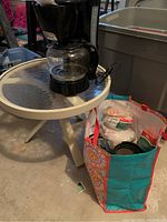 Westinghouse drip coffee maker on round outdoor table with reusable bag holding coffee accessories placed on floor next to table.