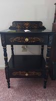 Front view of the black end table showing fruit and floral designs on upper backboard and drawer fronts, with a brass knob on each drawer.