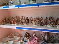 Wide shot of two shelves holding approximately 24 Hummel figurines and assorted ceramics