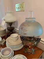 Two vintage hurricane lamps on a table with dishware in background, one with a white scalloped glass shade and the other with a greenish painted floral glass shade.
