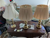 Three brass look table lamps positioned on a dark wood coffee table, showing two with pleated beige lampshades and one with a cream fabric shade with discoloration.