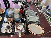 Top-down view of table with various kitchenware including Le Creuset and Fire King dishes, glass bowls, wooden-handled utensils, coasters, and hand grinder.