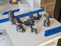 Seven miniature pewter and metal animal figures displayed on a white surface with blue tape markings showing approximate dimensions.