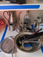 Wide view showing pewter teapot, cream jug, sugar bowl, two handled cup, glass bottle, bowl with utensils, candle snuffers, small figurines and metal plates.