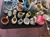 Wide view showing all pitchers in the lot on a black table. Includes various styles, colors, and sizes.
