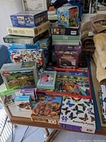 Multiple puzzles stacked on tables, shows puzzle boxes with varying pieces and artwork, arranged to show quantity and variety.