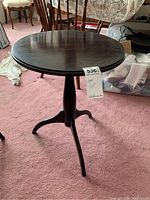 Front view of a dark finished round wooden table on a pink carpet, showing a central pedestal and three legs, with a tag attached to the base.