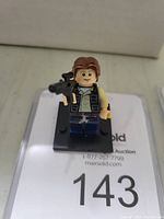 LEGO Han Solo mini-figure holding a blaster on a black stand with lot number 143 beneath.