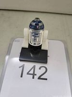 Photo of a LEGO R2-D2 mini-figure on a black base with lot number 142 on paper beneath it.