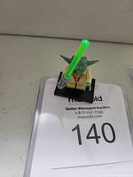LEGO Yoda mini-figure holding green lightsaber placed on black display stand with auction lot number card in background.
