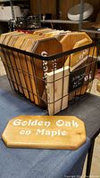 Photo of multiple wooden plaques stacked inside a black wire basket, showing edges and painted text on the plaques such as 'Golden Oak on Maple' and other handwritten notes.