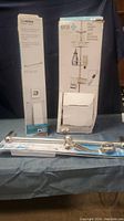 Photo showing boxed Moen Designer Grab Bar with Integrated Towel Bar and boxed Era Pole Shower Caddy with loose metal parts in front.