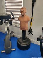 Tan mannequin torso punching bag on black base with exercise equipment around.