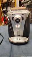 Front view of the Saeco coffee machine showing control knob with coffee cup icon, drip tray, and machine body.
