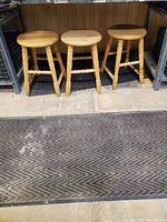 Three unfinished solid wood bar stools with round seats and turned legs, displayed side by side against a wooden backdrop and black shelving.