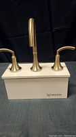 Front view showing the brushed gold finish Moen faucet set with three separate components mounted on a white box with Moen branding.