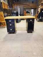 Front view of a solid wood desk showing the natural wood top and black base with three drawers and a side storage compartment.