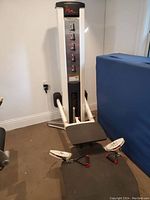 Full view of a white and black Step weight machine with foot pedals and padded areas, positioned against a wall.