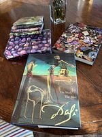 Photo of seven coffee table books stacked and spread on a wooden table, including Salvador Dali's surrealist artwork cover, Rolling Stones collage cover, and a book with purple grapes on the cover.