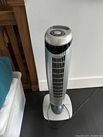 Full view of the silver standing oscillating tower fan showing the vent and base with power cord on the floor.
