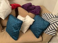Two teal throw pillows, white pillow cover, blue and white striped pillow cover, purple and red throws arranged on beige couch