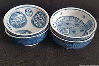 Side-by-side view of four bowls in two stacks, showing interior decoration of two with geometric patterns and two with fish motifs, with textured blue exterior.