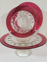 Front view showing clear glass pedestal cake stand with textured thumbprint design and two smaller side plates with ruby red rims and matching thumbprint glass pattern.
