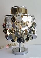 Full view of the chrome lamp with circular reflective metal disks hanging from the shade structure, showing the mixed silver and gold tones and two-tier design.