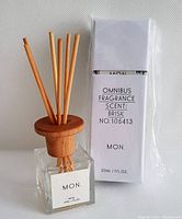 Glass square bottle with wooden top holding several reed diffuser sticks alongside original white box packaging labeled with scent 'Brisk' and product info.