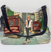 Front view of handbag showing cityscape design with woman and red cable car with beaded details, leather strap.