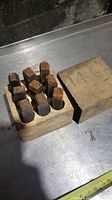 Wooden block with 11 metal punches arranged upright, showing their square tops and worn condition. Wooden lid labeled '146' rests beside them.