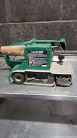 Side view of green 4 inch belt sander showing wooden handle, warning label, and metal base