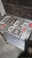 Top view of assortment of 14 C-clamps on metal table. Clamps vary in size from about 1 inch to 6 inches, with various metal finishes and paint colors on handles.